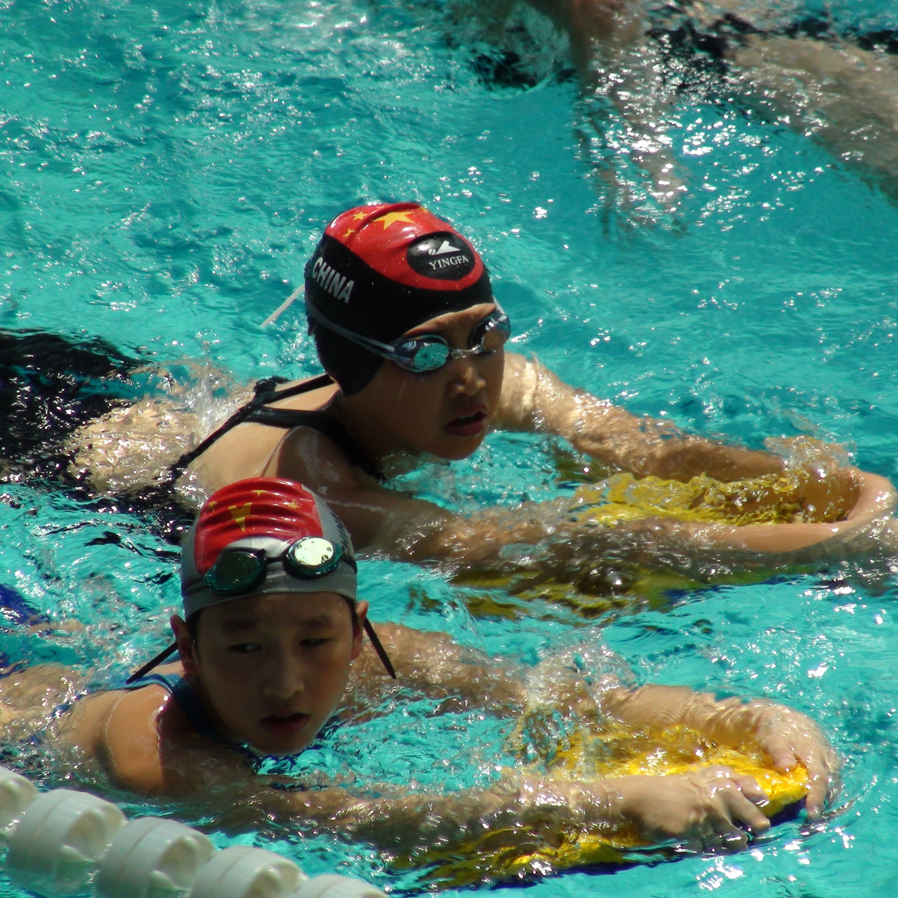 Young swimmers wearing caps and goggles practicing with kickboards in a pool